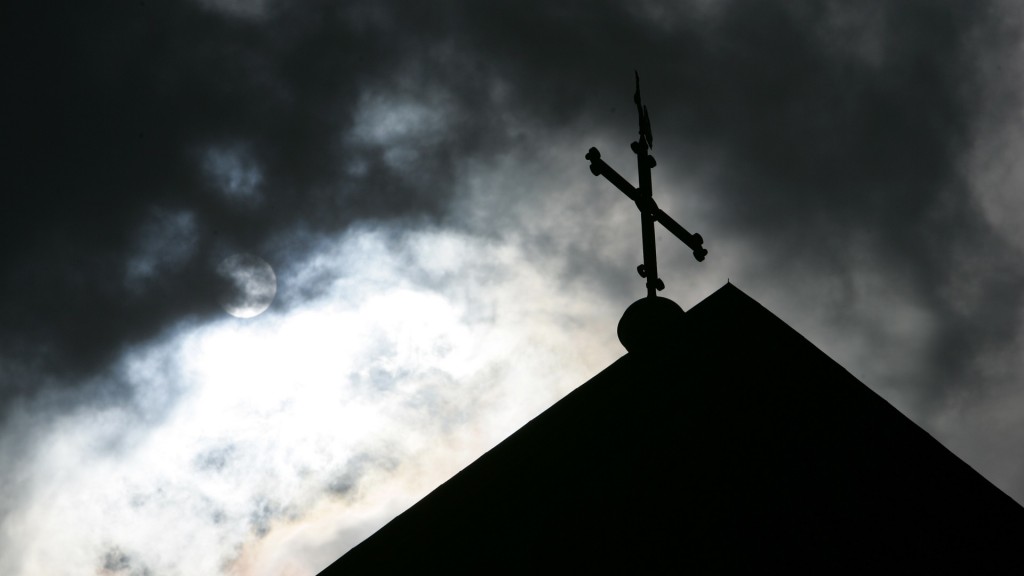 Im Gegenlicht und vor wolkenverhangenem Himmel ist die Kirchturmspitze mit Kreuz zu sehen (Foto: picture alliance/dpa | Friso Gentsch)