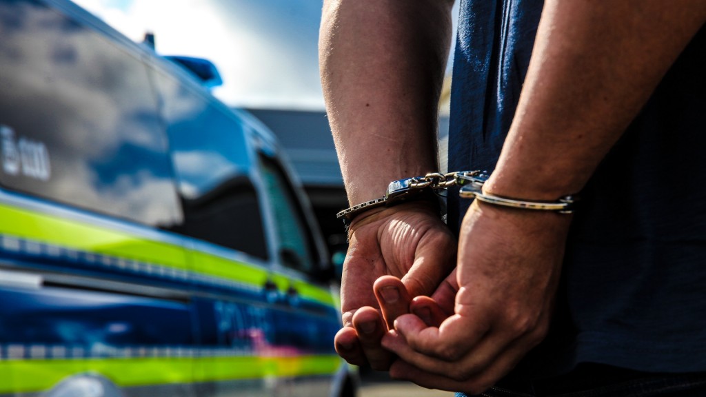 Foto: Eine Person wird durch die Polizei festgenommen, es wurden die Handschellen angelegt. Im Hintergrund sieht man den Streifenwagen der Polizei stehen.