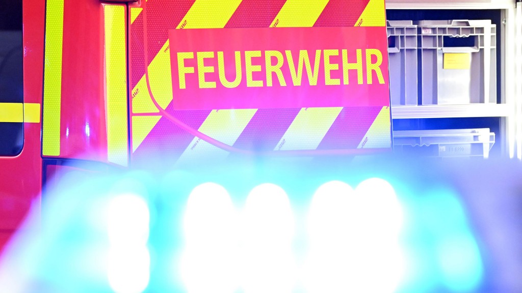 Ein Fahrzeug der Feuerwehr steht bei einem Einsatz auf einer Straße, während im Vordergrund das Blaulicht eines Polizeifahrzeugs zu sehen ist