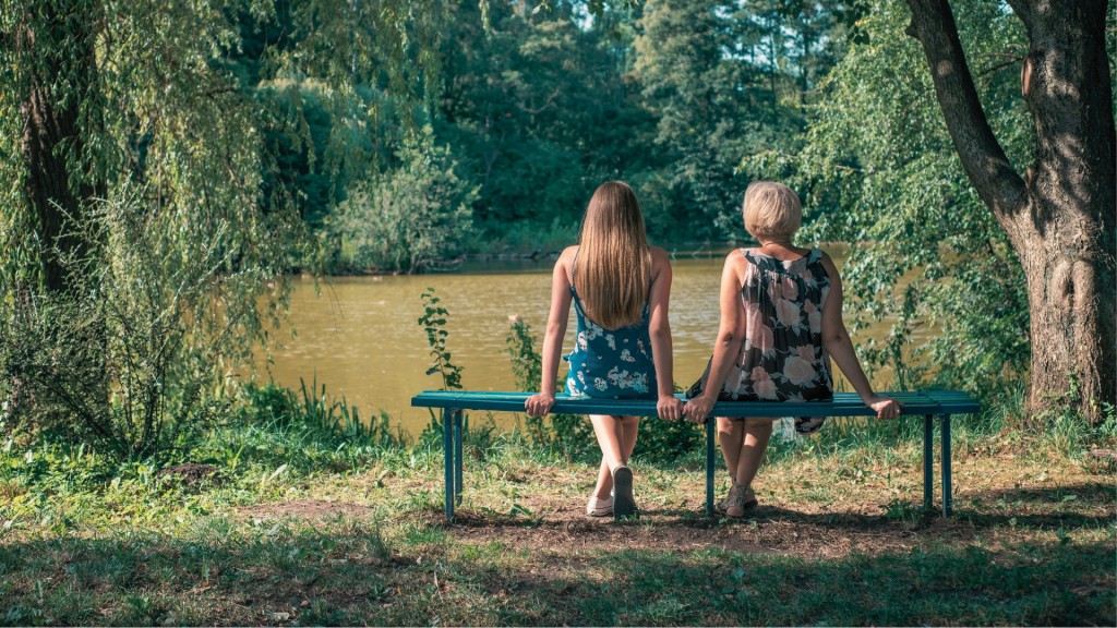 Das Bild oben zeigt eine alte Frau und eine Jugendliche von hinten, die nebeneinander auf einer Parkbank in der Natur sitzen.