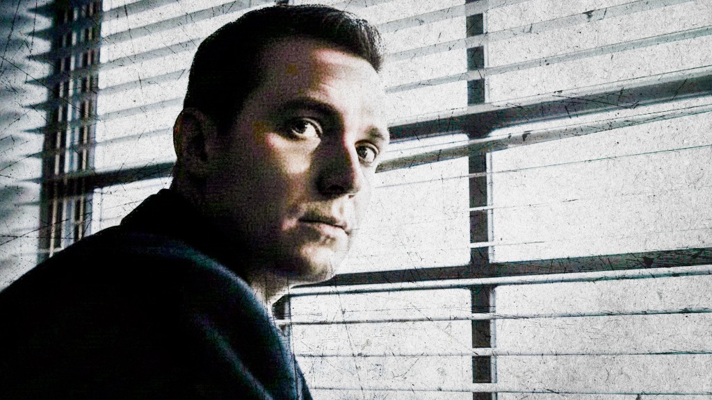 Ein Mann sitzt vor einem Fenster mit Jalousinen und schaut ernst in die Kamera. 