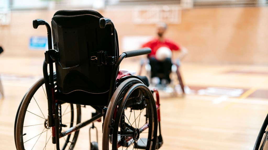 Ein leerer Rollstuhl steht in einer Sporthalle, im Hintergrund ein Rollstuhlfahrer mit einem Spielball
