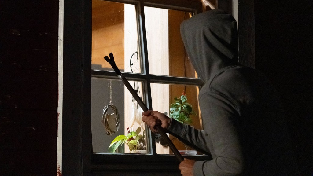 Symbolbild: Ein Einbrecher mit Kapuze und Brecheisen steht nachts vor einem gekippten Fenster