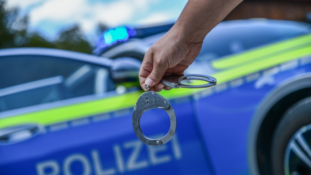 Foto: Eine Person hält Handschellen in der Hand. Im Hintergrund steht ein Polizeifahrzeug