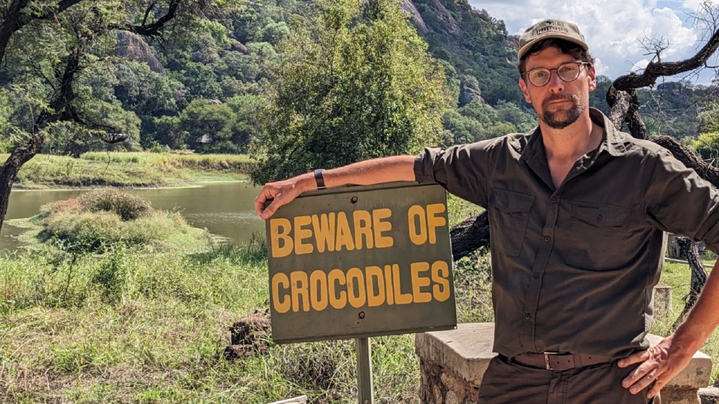 Christoph Borgans steht in Afrika neben einem Schild mit der Aufschrift 'Beware of Crocodiles'.