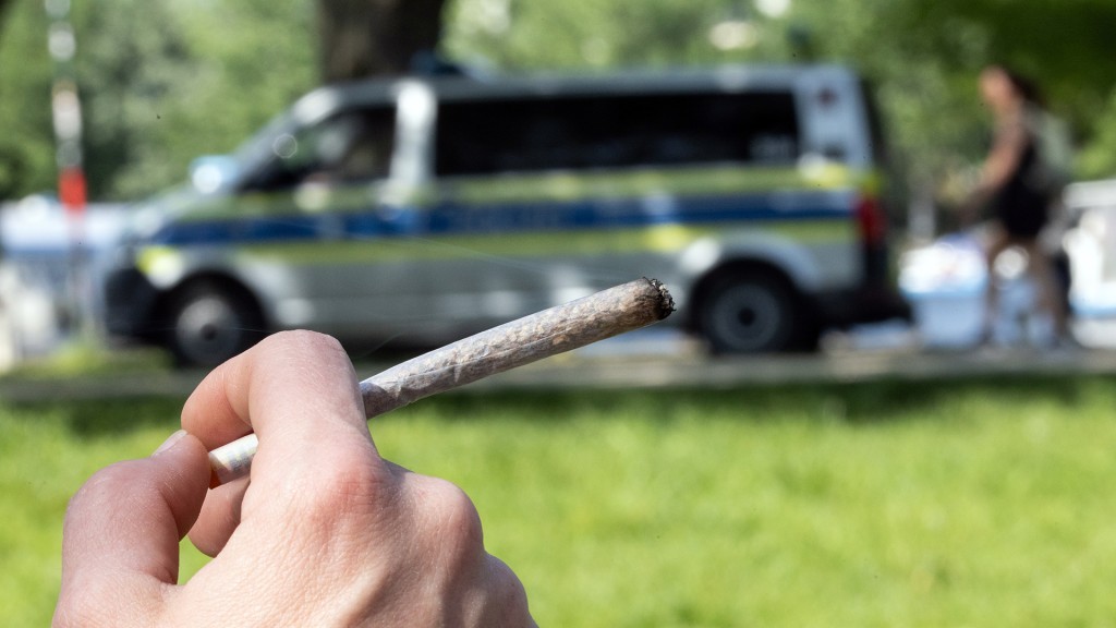 Jemand hält einen Joint in der Hand, im Hintergrund sieht man ein Polizeifahrzeug