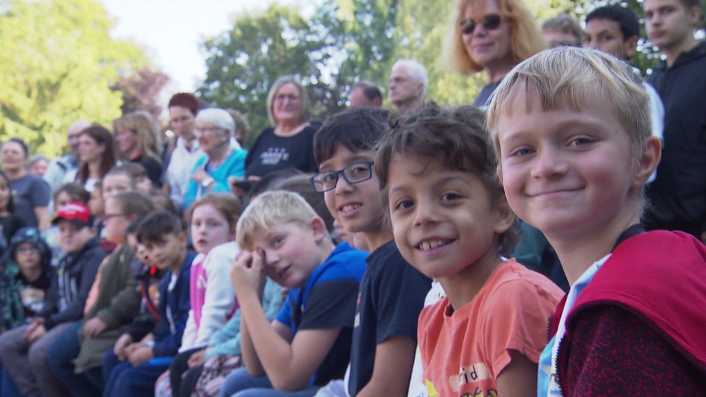 Foto: Kinder schauen in die Kamera und Erwachsene im Hintergrund