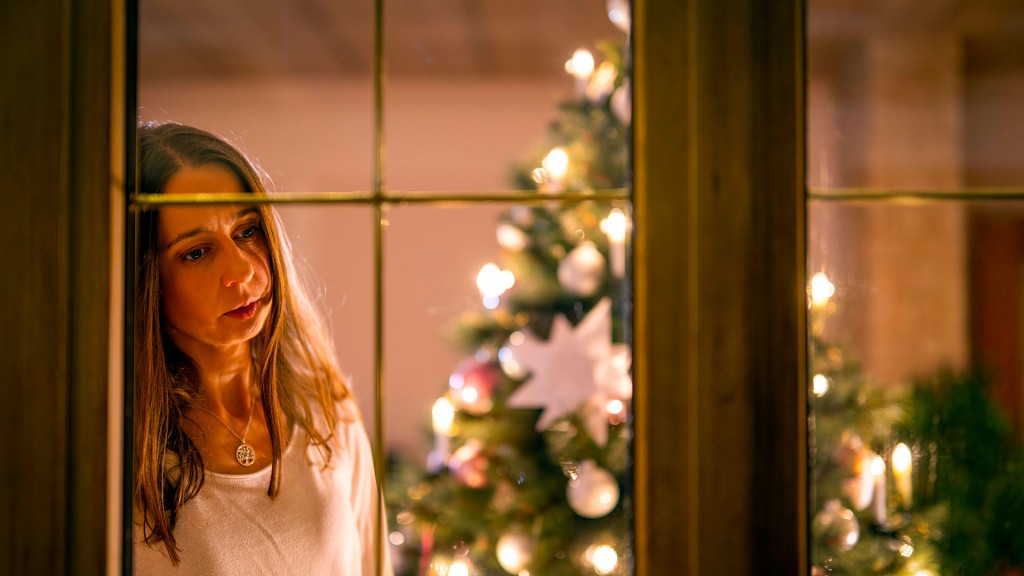 Eine Frau schaut nachdenklich aus dem Wohnzimmer durchs Fenster, dahinter ein festlich dekorierter Weihnachtsbaum