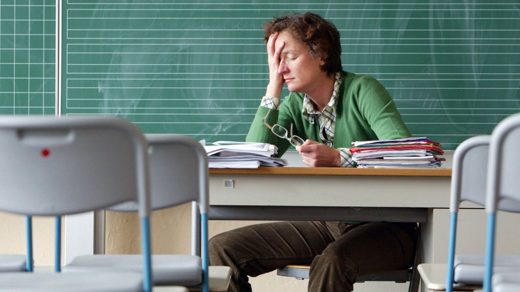 Foto: Jemand sitzt mit zahlreichen Unterlagen an einem Lehrerpult vor der Tafel und stützt das Gesicht in die Hand