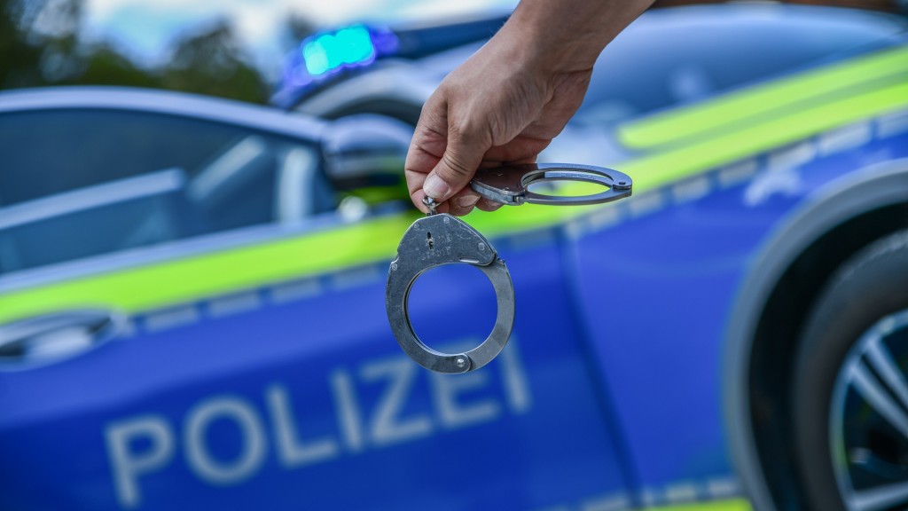 Foto: Eine Person hält Handschellen in der Hand. Im Hintergrund steht ein Polizeifahrzeug