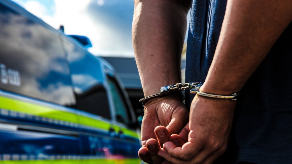 Symbolbild: Eine Person wird durch die Polizei festgenommen, es wurden die Handschellen angelegt. Im Hintergrund sieht man den Streifenwagen der Polizei stehen.